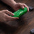 A person's hands holding a translucent green retro game controller over a dark wood table, with a black game controller and two small black accessories nearby.