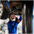 A mechanic in a blue uniform works underneath a raised car using a red power tool in an auto repair shop.