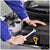 A close-up of a mechanic in a blue uniform working under a car hood, using a wrench near a yellow oil dipstick.