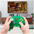 A person holds a translucent green retro-style game controller for the Nintendo 64/N64 in front of a TV showing a fighting game scene. The controller has a white analog stick, a gray D-pad, yellow action buttons, and a red button, with a black cable connected.